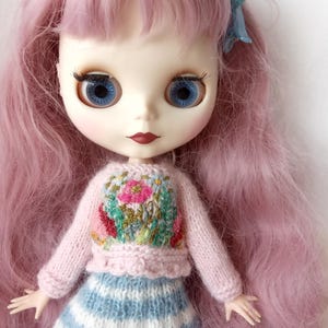 K&ouml;nnte beinhalten: Blythe-Puppe mit langen rosa Haaren und blauen Augen. Die Puppe tr&auml;gt einen hellrosa Pullover mit gesticktem Blumenmuster und einen blau-wei&szlig; gestreiften Rock. Eine blaue Schleife schm&uuml;ckt das Haar.