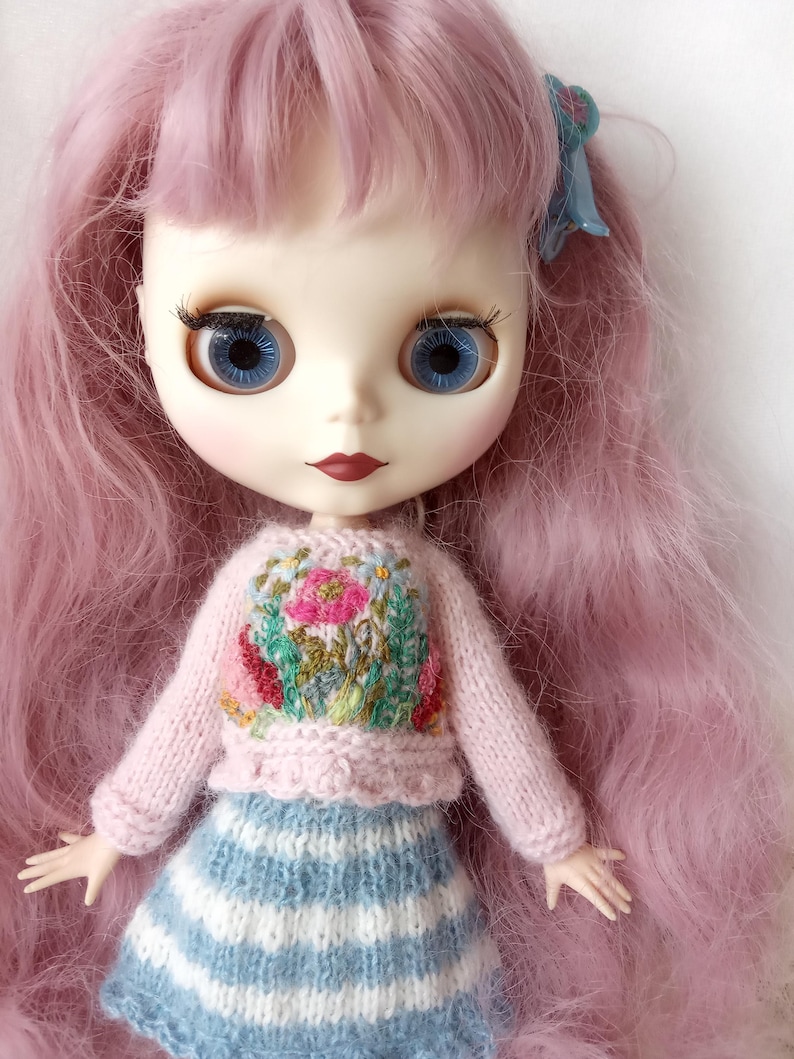 K&ouml;nnte beinhalten: Blythe-Puppe mit langen rosa Haaren und blauen Augen. Die Puppe tr&auml;gt einen hellrosa Pullover mit gesticktem Blumenmuster und einen blau-wei&szlig; gestreiften Rock. Eine blaue Schleife schm&uuml;ckt das Haar.