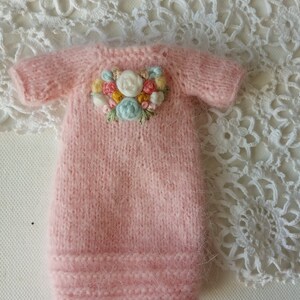 Puede incluir: Un vestido de beb&eacute; de punto rosa con un dise&ntilde;o de bordado floral en la parte delantera.