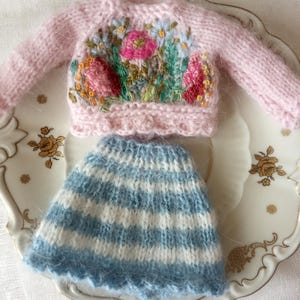 K&ouml;nnte beinhalten: Ein Miniatur-Set aus einem rosa Strickpullover mit Blumenstickerei und einem blau-wei&szlig; gestreiften Rock. Der Pullover hat einen Rundhalsausschnitt und lange &Auml;rmel. Der Rock hat einen gewellten Saum. Das Set wird auf einem dekorativen Teller pr&auml;sentiert.