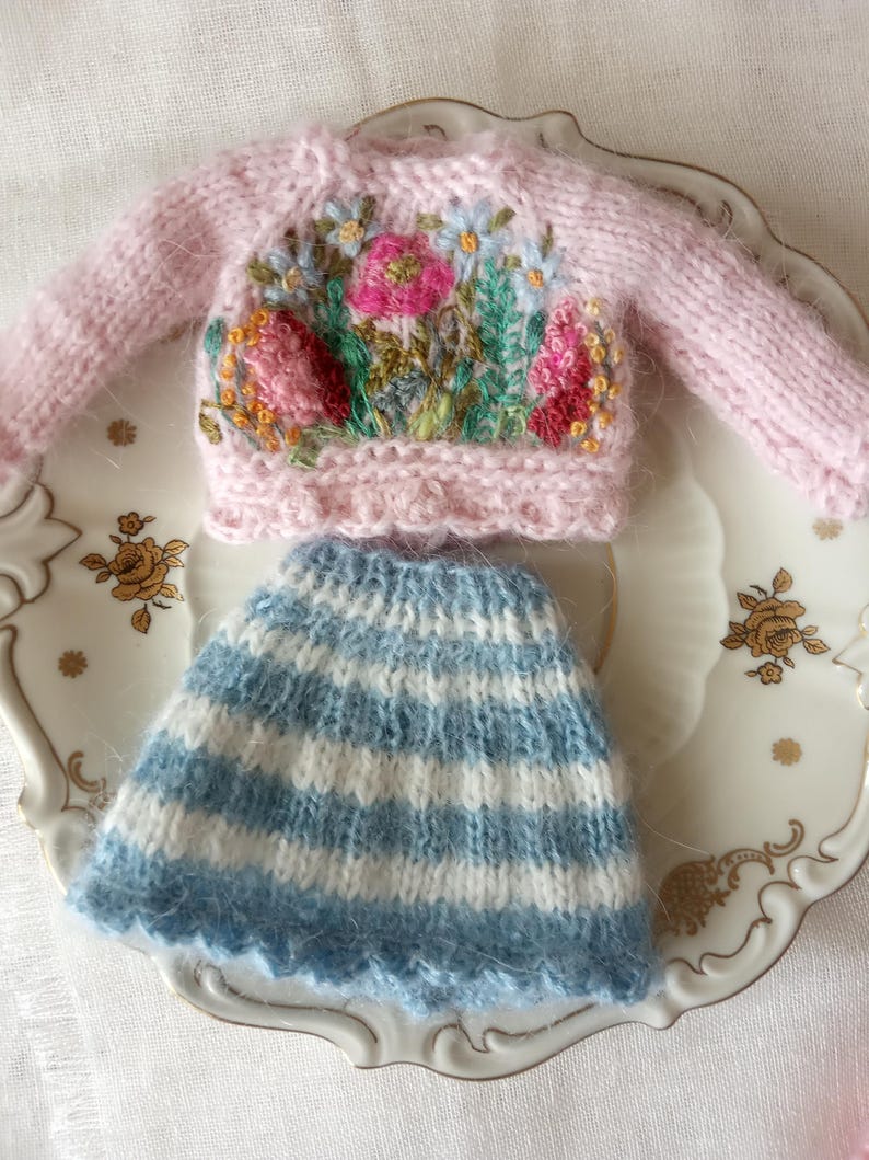 K&ouml;nnte beinhalten: Ein Miniatur-Set aus einem rosa Strickpullover mit Blumenstickerei und einem blau-wei&szlig; gestreiften Rock. Der Pullover hat einen Rundhalsausschnitt und lange &Auml;rmel. Der Rock hat einen gewellten Saum. Das Set wird auf einem dekorativen Teller pr&auml;sentiert.