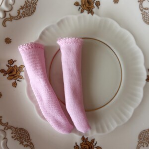 K&ouml;nnte beinhalten: Zwei Paar hellrosa Puppensocken liegen auf einem wei&szlig;en Teller mit goldenem Blumenmuster. Die Socken sind aus weichem Stoff und haben einen ger&uuml;schten Rand. Der Teller hat einen gewellten Rand.
