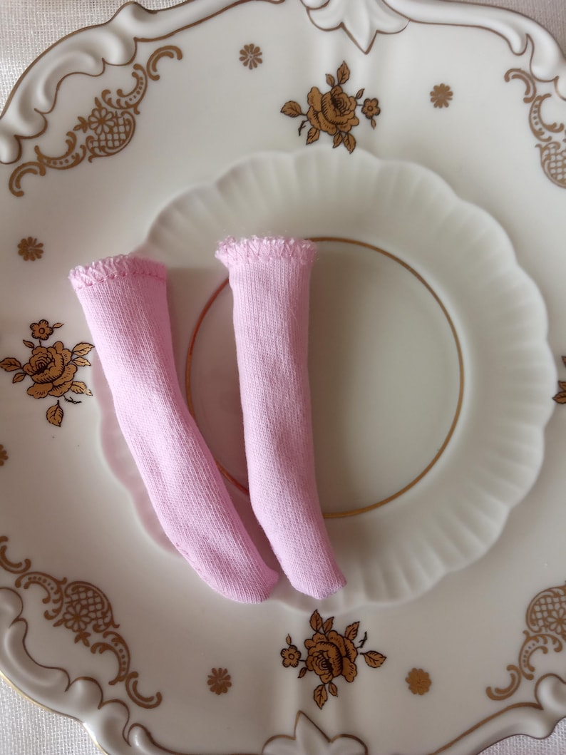 K&ouml;nnte beinhalten: Zwei Paar hellrosa Puppensocken liegen auf einem wei&szlig;en Teller mit goldenem Blumenmuster. Die Socken sind aus weichem Stoff und haben einen ger&uuml;schten Rand. Der Teller hat einen gewellten Rand.