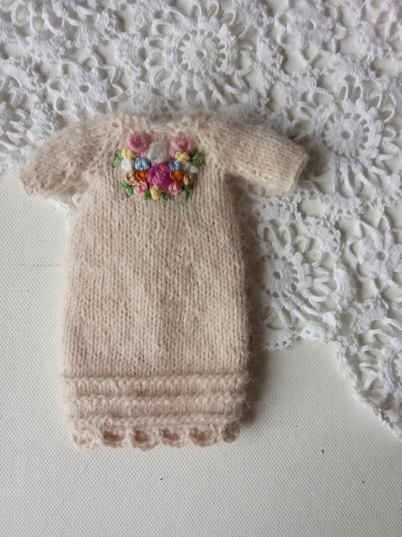 Puede incluir: Un vestido de beb&eacute; de punto beige con un dise&ntilde;o de bordado floral en el escote.