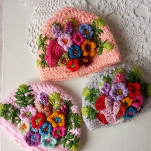 Könnte beinhalten: Drei gehäkelte Mützen mit bunten gestickten Blumen. Die Mützen sind rosa, grau und rosa mit grünen Akzenten.