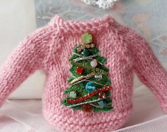 May include: A pink knitted sweater with a green embroidered Christmas tree decorated with silver tinsel, gold beads, and colorful rhinestones.