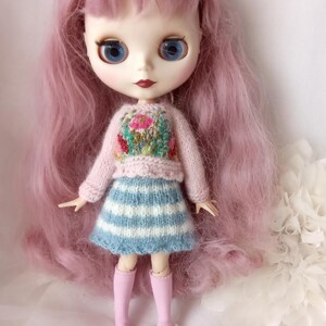 K&ouml;nnte beinhalten: Eine Puppe mit langen rosa Haaren, blauen Augen und roten Lippen. Die Puppe tr&auml;gt einen rosa Pullover mit floralem Design, einen blau-wei&szlig; gestreiften Rock und rosa kniehohe Socken. Eine kleine blaue Schleife befindet sich im Haar der Puppe.