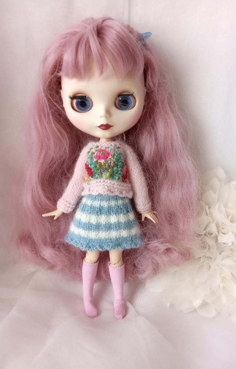 K&ouml;nnte beinhalten: Eine Puppe mit langen rosa Haaren, blauen Augen und roten Lippen. Die Puppe tr&auml;gt einen rosa Pullover mit floralem Design, einen blau-wei&szlig; gestreiften Rock und rosa kniehohe Socken. Eine kleine blaue Schleife befindet sich im Haar der Puppe.