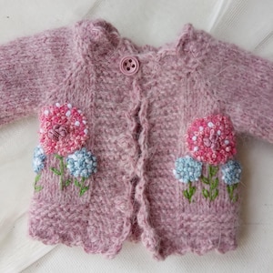 May include: A pink knitted baby cardigan with embroidered flowers in shades of pink, blue, and green. The cardigan has a button closure and a scalloped edge.
