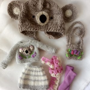 May include: A collection of handmade knitted doll clothes, including a koala bear hat, sweater, skirt, scarf, purse, and leggings. The hat and sweater feature koala bear faces. The skirt is striped, and the scarf is pink and white.