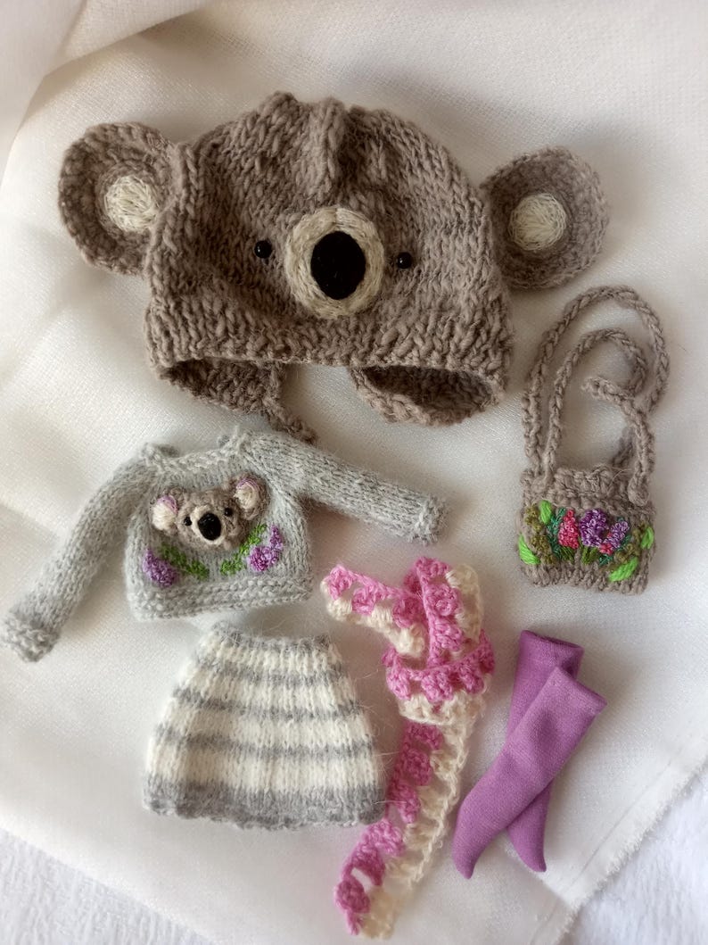 May include: A collection of handmade knitted doll clothes, including a koala bear hat, sweater, skirt, scarf, purse, and leggings. The hat and sweater feature koala bear faces. The skirt is striped, and the scarf is pink and white.