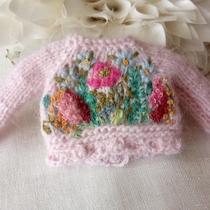 K&ouml;nnte beinhalten: Ein hellrosa, miniatur Strickpullover mit langen &Auml;rmeln. Der Pullover hat ein gesticktes Blumenmuster mit rosa, roten, blauen und gelben Blumen und gr&uuml;nen Bl&auml;ttern. Der untere Saum hat einen gewellten Rand.