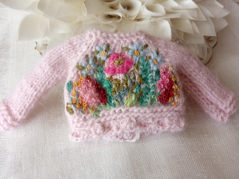 K&ouml;nnte beinhalten: Ein hellrosa, miniatur Strickpullover mit langen &Auml;rmeln. Der Pullover hat ein gesticktes Blumenmuster mit rosa, roten, blauen und gelben Blumen und gr&uuml;nen Bl&auml;ttern. Der untere Saum hat einen gewellten Rand.