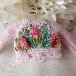 K&ouml;nnte beinhalten: Ein kleiner, rosa gestrickter Pullover mit floralen Stickereien. Der Pullover hat einen Rundhalsausschnitt und lange &Auml;rmel. Die Stickerei zeigt rosa, rote, blaue und gelbe Blumen mit gr&uuml;nen Bl&auml;ttern. Der Pullover liegt auf einer wei&szlig;en Oberfl&auml;che.