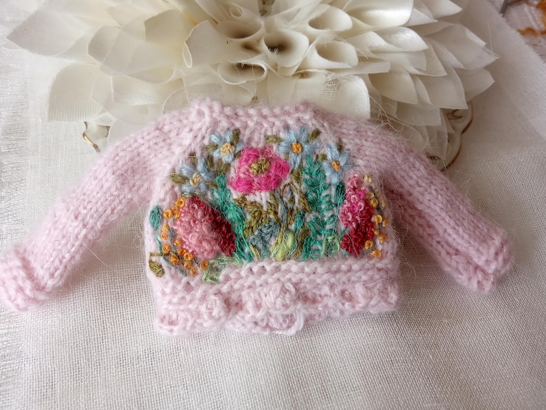 K&ouml;nnte beinhalten: Ein kleiner, rosa gestrickter Pullover mit floralen Stickereien. Der Pullover hat einen Rundhalsausschnitt und lange &Auml;rmel. Die Stickerei zeigt rosa, rote, blaue und gelbe Blumen mit gr&uuml;nen Bl&auml;ttern. Der Pullover liegt auf einer wei&szlig;en Oberfl&auml;che.