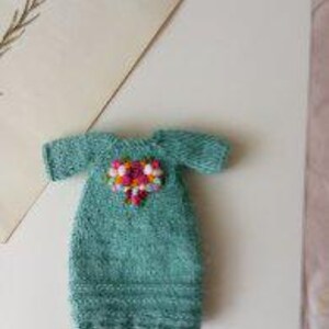 Puede incluir: Un vestido de punto turquesa para mu&ntilde;eca con un adorno floral rosa y blanco en la parte delantera.