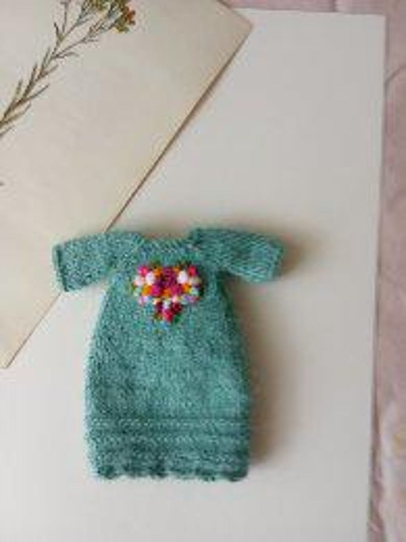 Puede incluir: Un vestido de punto turquesa para mu&ntilde;eca con un adorno floral rosa y blanco en la parte delantera.