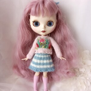 K&ouml;nnte beinhalten: Eine Puppe mit langen, rosa Haaren und gro&szlig;en blauen Augen. Die Puppe tr&auml;gt einen hellrosa Pullover mit Blumenstickerei, einen blau-wei&szlig; gestreiften Rock und rosa kniehohe Stiefel. Das Gesicht der Puppe ist mit roten Lippen bemalt.