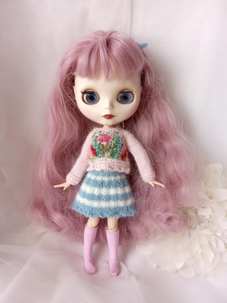 K&ouml;nnte beinhalten: Eine Puppe mit langen, rosa Haaren und gro&szlig;en blauen Augen. Die Puppe tr&auml;gt einen hellrosa Pullover mit Blumenstickerei, einen blau-wei&szlig; gestreiften Rock und rosa kniehohe Stiefel. Das Gesicht der Puppe ist mit roten Lippen bemalt.