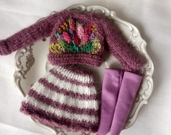 Könnte beinhalten: Ein Miniatur-Strick-Outfit in Lila- und Weißtönen, bestehend aus einem Pullover mit Blumenstickerei, einem gestreiften Rock und einer Leggings. Das Outfit wird auf einem dekorativen Teller präsentiert, der die handgefertigten Details hervorhebt.