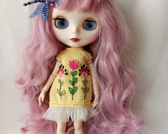 May include: A doll with pink curly hair wearing a yellow knitted dress with embroidered flowers and a blue dragonfly hair clip.