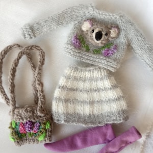 May include: A miniature knitted outfit for a doll, featuring a gray sweater with a koala face and purple flower accents, a striped skirt, a small purse with floral embroidery, and purple leggings. The outfit is designed for a doll.