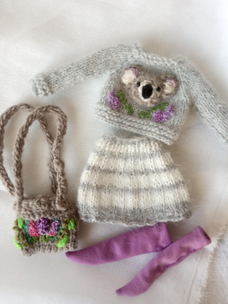 May include: A miniature knitted outfit for a doll, featuring a gray sweater with a koala face and purple flower accents, a striped skirt, a small purse with floral embroidery, and purple leggings. The outfit is designed for a doll.