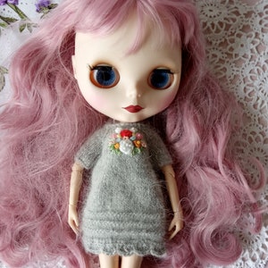 Puede incluir: Una mu&ntilde;eca con el pelo largo de color rosa que lleva un vestido de punto gris con flores bordadas. La mu&ntilde;eca tiene ojos azules y labios rojos.