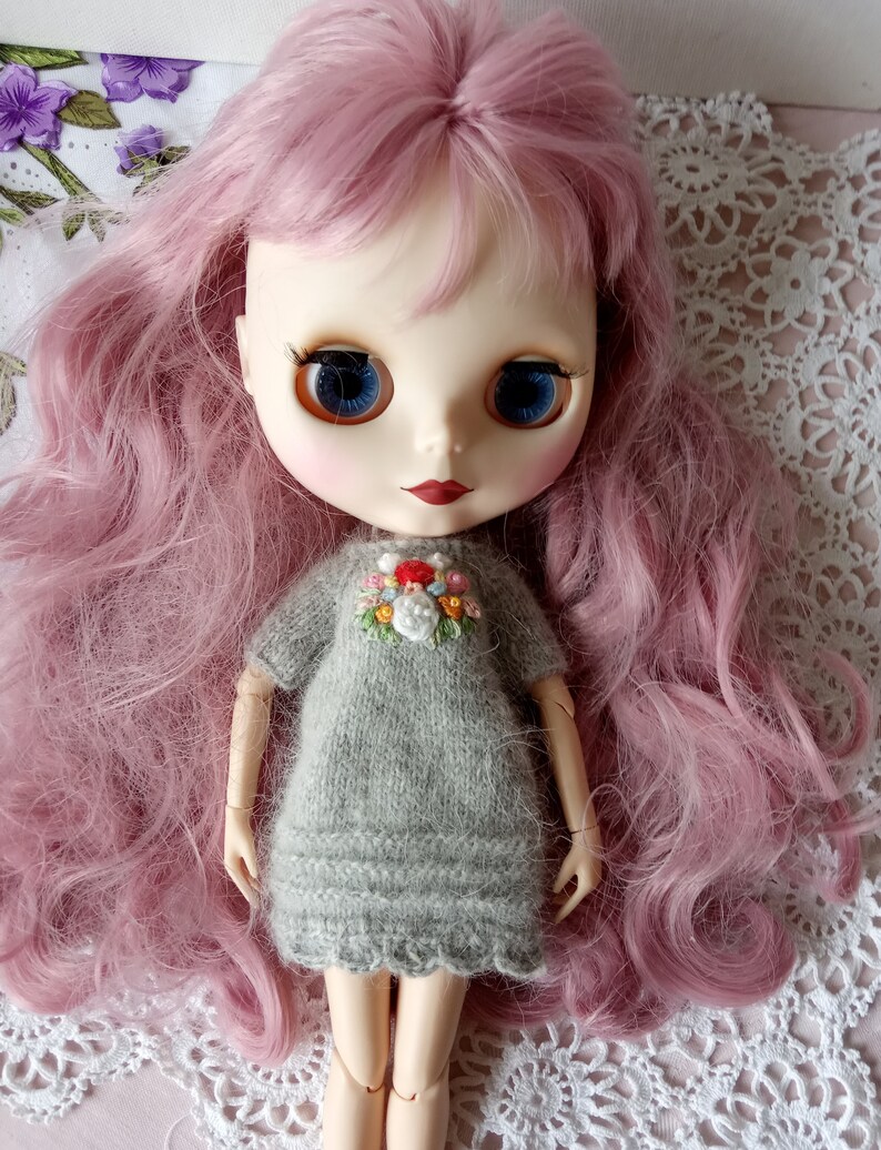 Puede incluir: Una mu&ntilde;eca con el pelo largo de color rosa que lleva un vestido de punto gris con flores bordadas. La mu&ntilde;eca tiene ojos azules y labios rojos.