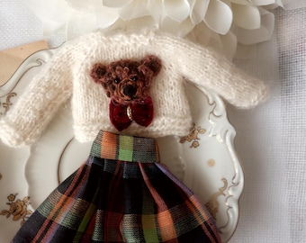 Könnte beinhalten: Ein Miniatur-Outfit für eine Puppe, bestehend aus einem cremefarbenen Strickpullover mit einem braunen Teddybär-Applikation und einer roten Fliege. Das Outfit beinhaltet einen karierten Rock in Schwarz-, Orange-, Grün- und Lilatönen.