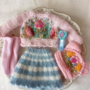 K&ouml;nnte beinhalten: Ein Miniatur-Puppenkleidungsset mit einem rosa Pullover mit Blumenstickerei, einem blau-wei&szlig; gestreiften Rock, rosa Socken und einem passenden Hut. Ein blaues herzf&ouml;rmiges Accessoire ist ebenfalls enthalten. Das Set wird auf einem dekorativen Teller pr&auml;sentiert.