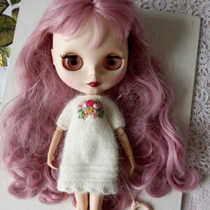 Puede incluir: Una mu&ntilde;eca con el pelo rosa que lleva un vestido blanco de punto con flores bordadas. La mu&ntilde;eca tiene ojos marrones y labios rojos.