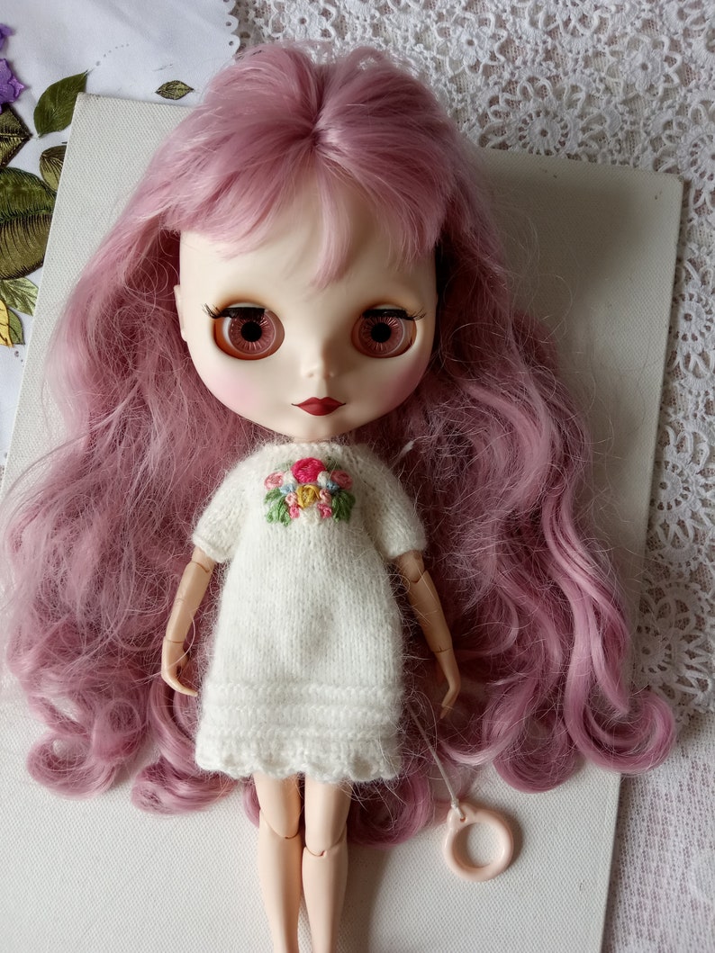 Puede incluir: Una mu&ntilde;eca con el pelo rosa que lleva un vestido blanco de punto con flores bordadas. La mu&ntilde;eca tiene ojos marrones y labios rojos.