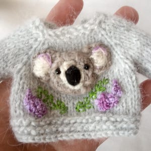 May include: A miniature gray knit sweater with a koala face and purple flowers embroidered on the front. The koala has a black nose and eyes, with pink accents on the ears. The sweater is held in a hand.