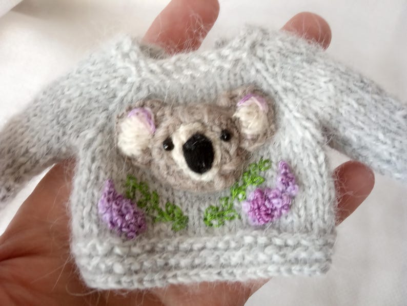May include: A miniature gray knit sweater with a koala face and purple flowers embroidered on the front. The koala has a black nose and eyes, with pink accents on the ears. The sweater is held in a hand.