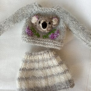 May include: A miniature gray knit sweater and skirt set. The sweater features a koala face with purple flowers and green leaves. The skirt has horizontal stripes of gray and white. The set is designed for a doll or small toy.