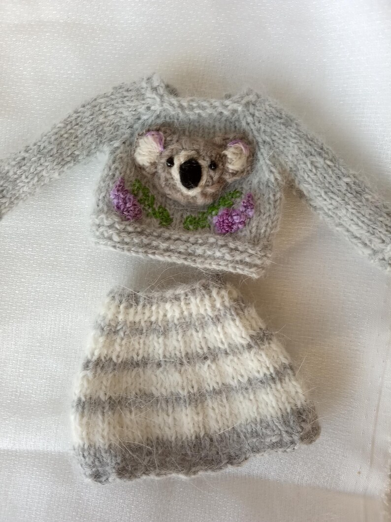 May include: A miniature gray knit sweater and skirt set. The sweater features a koala face with purple flowers and green leaves. The skirt has horizontal stripes of gray and white. The set is designed for a doll or small toy.
