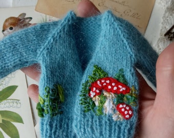 Könnte beinhalten: Eine hellblaue Miniatur-Strickjacke mit detaillierter Stickerei aus rot-weiß gepunkteten Pilzen und grünem Blattwerk. Das Kleidungsstück hat lange Ärmel, einen gerippten Saum und eine weiche, flauschige Textur mit feiner Handarbeit.