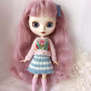 K&ouml;nnte beinhalten: Puppe mit langen rosa Haaren, blauen Augen und hellem Teint. Die Puppe tr&auml;gt einen rosa Pullover mit Blumenstickerei, einen blau-wei&szlig; gestreiften Rock und rosa kniehohe Socken. Eine blaue Schleife schm&uuml;ckt das Haar der Puppe.
