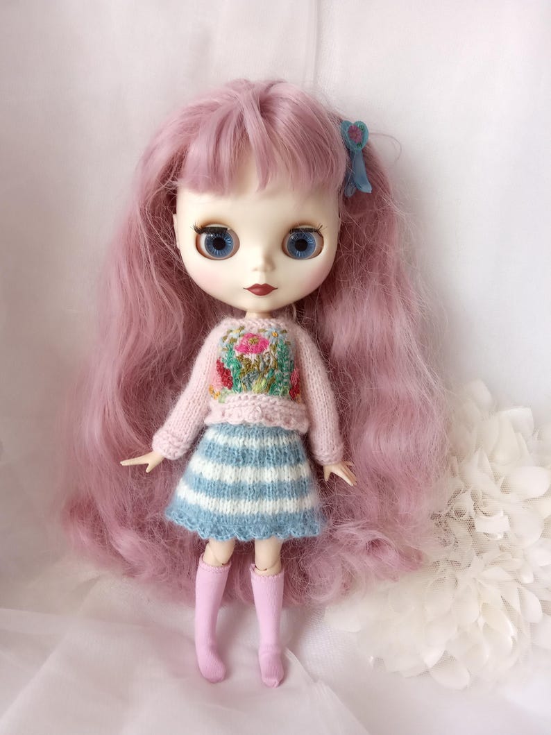 K&ouml;nnte beinhalten: Puppe mit langen rosa Haaren, blauen Augen und hellem Teint. Die Puppe tr&auml;gt einen rosa Pullover mit Blumenstickerei, einen blau-wei&szlig; gestreiften Rock und rosa kniehohe Socken. Eine blaue Schleife schm&uuml;ckt das Haar der Puppe.