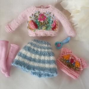 K&ouml;nnte beinhalten: Eine Sammlung von Miniatur-Puppenkleidung, darunter ein rosa Pullover mit Blumenstickerei, ein blau-wei&szlig; gestreifter Rock, rosa Socken und ein rosa Oberteil mit Blumenstickerei. Eine blaue Haarspange mit herzf&ouml;rmiger Blume ist ebenfalls enthalten.