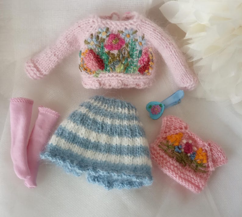 K&ouml;nnte beinhalten: Eine Sammlung von Miniatur-Puppenkleidung, darunter ein rosa Pullover mit Blumenstickerei, ein blau-wei&szlig; gestreifter Rock, rosa Socken und ein rosa Oberteil mit Blumenstickerei. Eine blaue Haarspange mit herzf&ouml;rmiger Blume ist ebenfalls enthalten.
