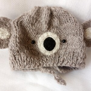 May include: A knitted gray koala bear hat with attached ears and a face. The hat features a white and black face with small black bead eyes. The hat has a tie under the chin.