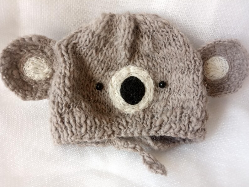 May include: A knitted gray koala bear hat with attached ears and a face. The hat features a white and black face with small black bead eyes. The hat has a tie under the chin.