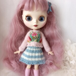 K&ouml;nnte beinhalten: Puppe mit langen, rosa Haaren und blauen Augen. Die Puppe tr&auml;gt einen hellrosa Pullover mit floralem Design, einen blau-wei&szlig; gestreiften Rock und rosa Stiefel. Eine blaue Schleife schm&uuml;ckt das Haar der Puppe.
