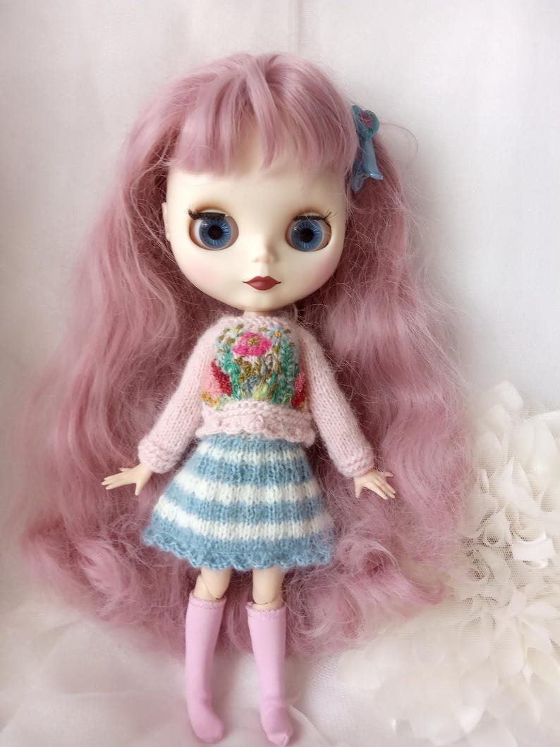 K&ouml;nnte beinhalten: Puppe mit langen, rosa Haaren und blauen Augen. Die Puppe tr&auml;gt einen hellrosa Pullover mit floralem Design, einen blau-wei&szlig; gestreiften Rock und rosa Stiefel. Eine blaue Schleife schm&uuml;ckt das Haar der Puppe.