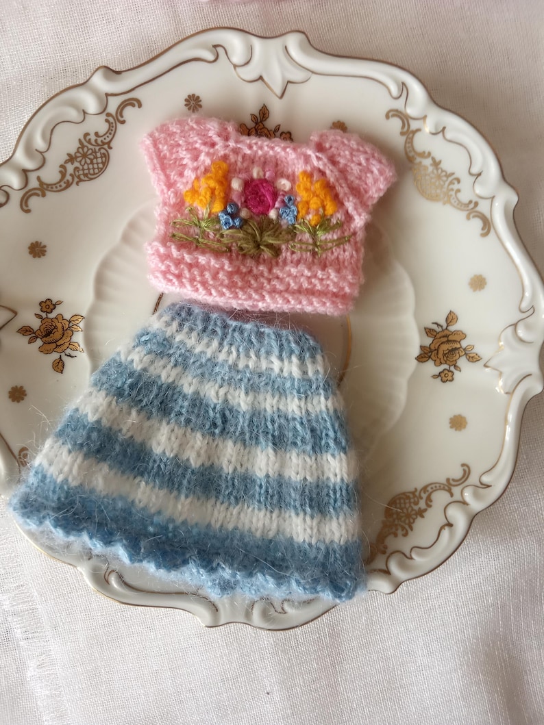 K&ouml;nnte beinhalten: Ein gestricktes Miniatur-Outfit, bestehend aus einem rosa Oberteil mit Blumenstickerei und einem blau-wei&szlig; gestreiften Rock. Das Outfit wird auf einem dekorativen wei&szlig;en Teller mit goldenen Akzenten pr&auml;sentiert, m&ouml;glicherweise f&uuml;r eine Puppe oder ein Bastelprojekt.
