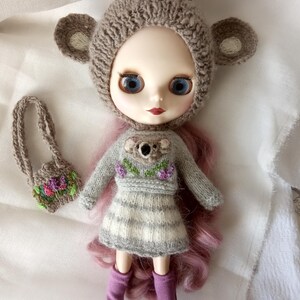 May include: A doll wearing a gray knitted koala bear hat and matching sweater with a koala applique and floral embroidery. The doll has blue eyes, pink hair, and purple boots. A small purse with floral embroidery is also included.