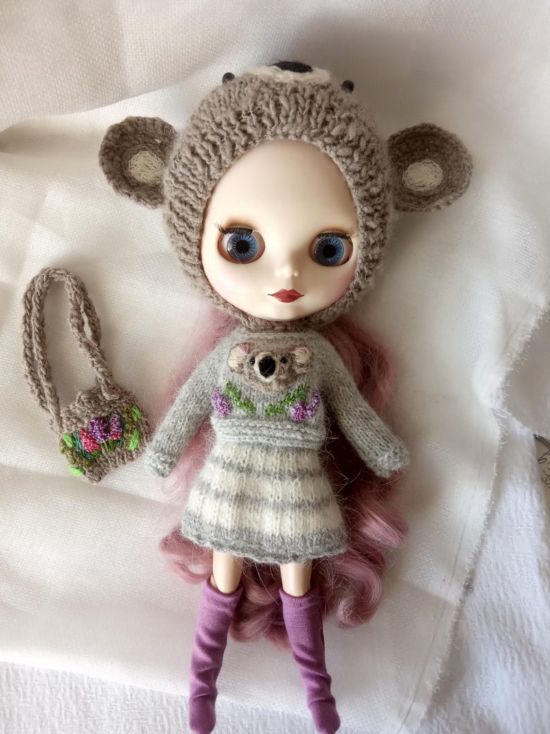 May include: A doll wearing a gray knitted koala bear hat and matching sweater with a koala applique and floral embroidery. The doll has blue eyes, pink hair, and purple boots. A small purse with floral embroidery is also included.