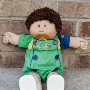 May include: A Cabbage Patch Kid doll with brown hair, wearing a green and yellow jumpsuit with blue trim and a pacifier. The doll is sitting on a concrete surface.